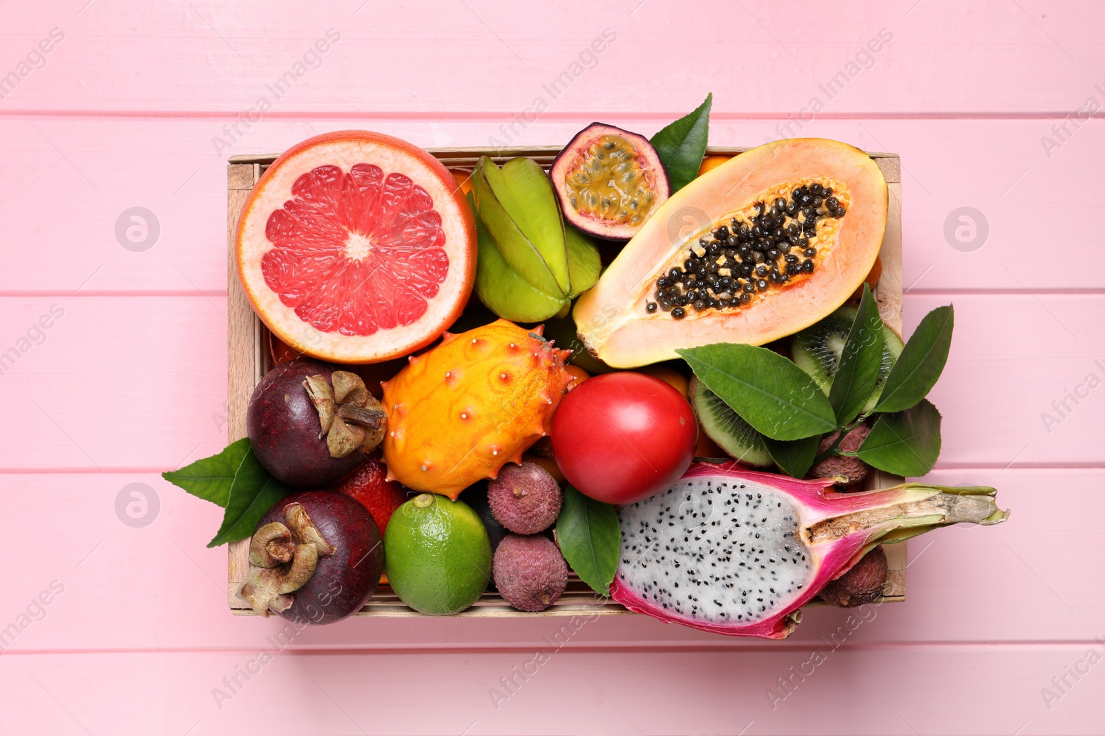 Photo of Different tropical fruits in box on pink wooden background, top view