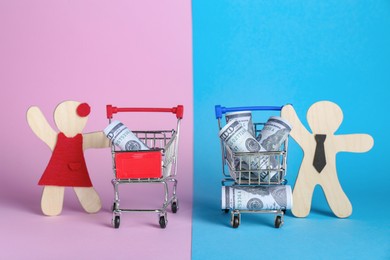 Photo of Gender pay gap. Wooden figures of man and woman near shopping carts with banknotes on color background