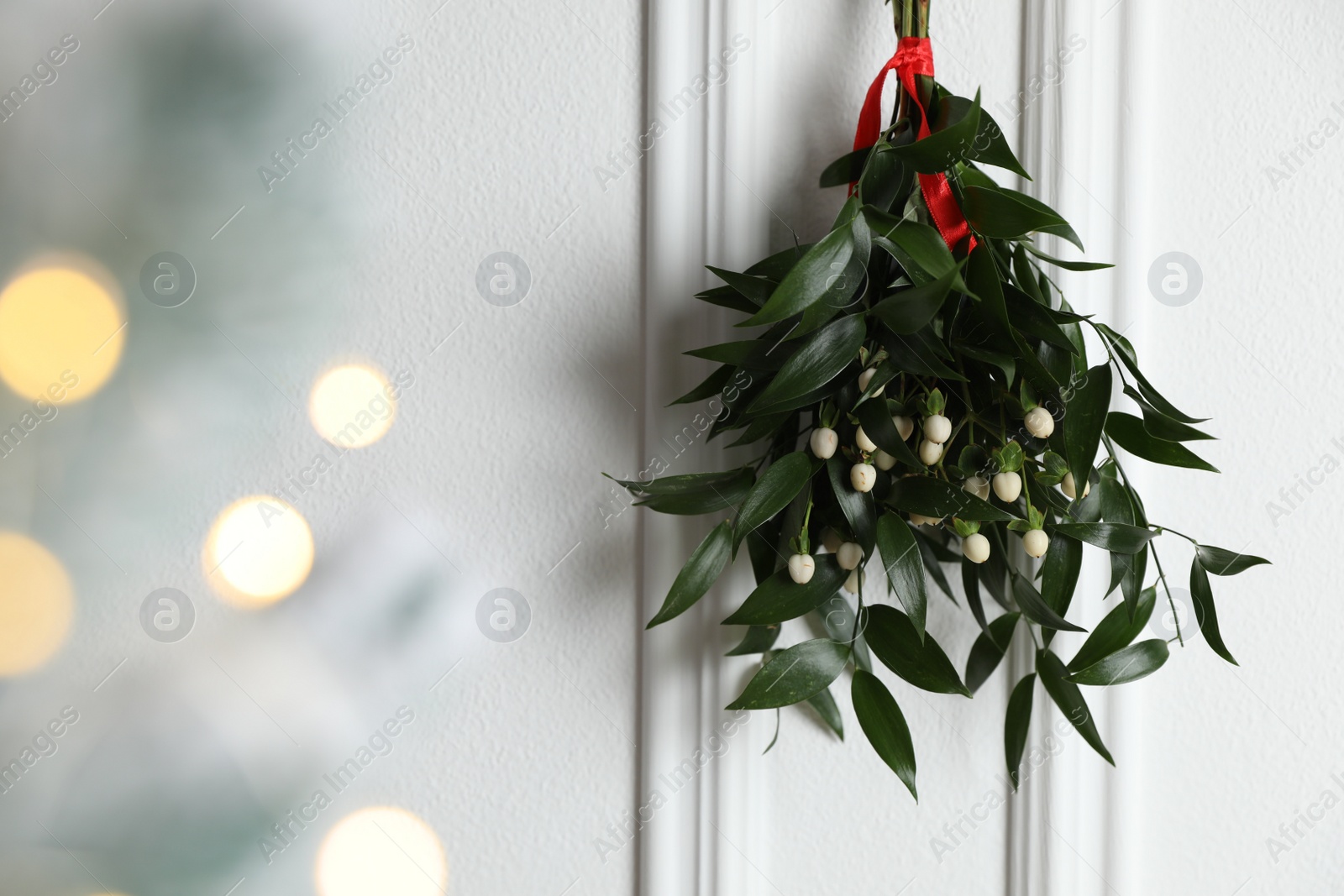 Photo of Mistletoe bunch with red ribbon hanging on light background, space for text. Bokeh effect