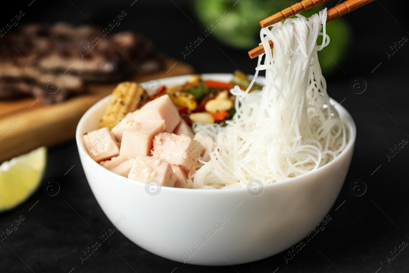 Photo of Chopsticks with tasty cooked rice noodles over bowl on black table, closeup