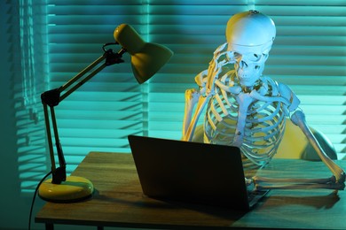 Photo of Waiting concept. Human skeleton sitting at wooden table with laptop indoors