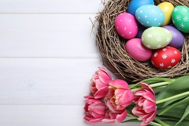 Colorful eggs, nest and tulips on white wooden background, flat lay with space for text. Happy Easter