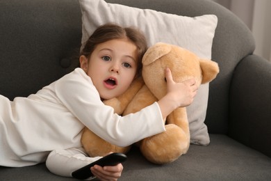 Little girl with remote control covering eyes to her toy bear while watching TV on sofa at home