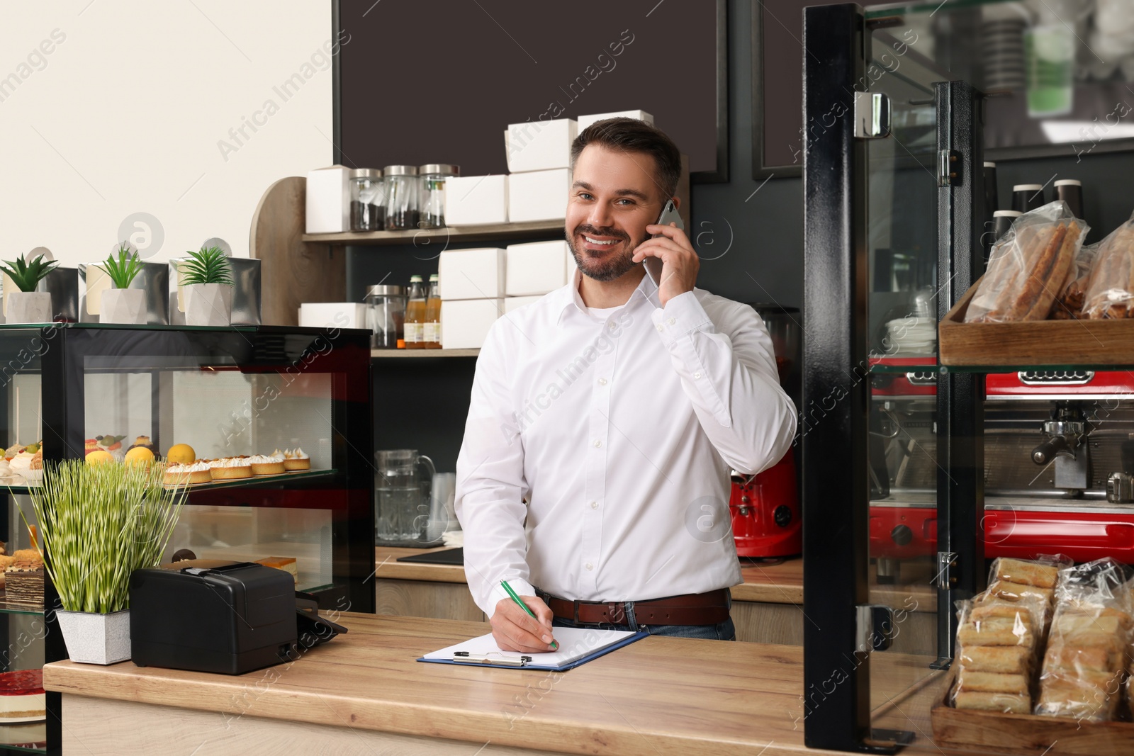 Photo of Business owner in his cafe. Man with clipboard talking on phone near showcases with pastries