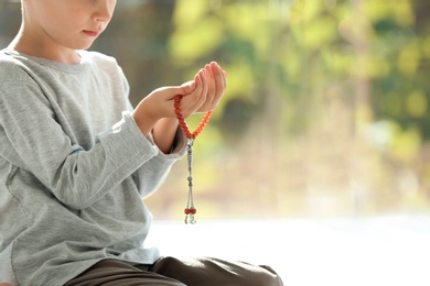 Photo of Little Muslim boy with misbaha praying indoors, closeup. Space for text