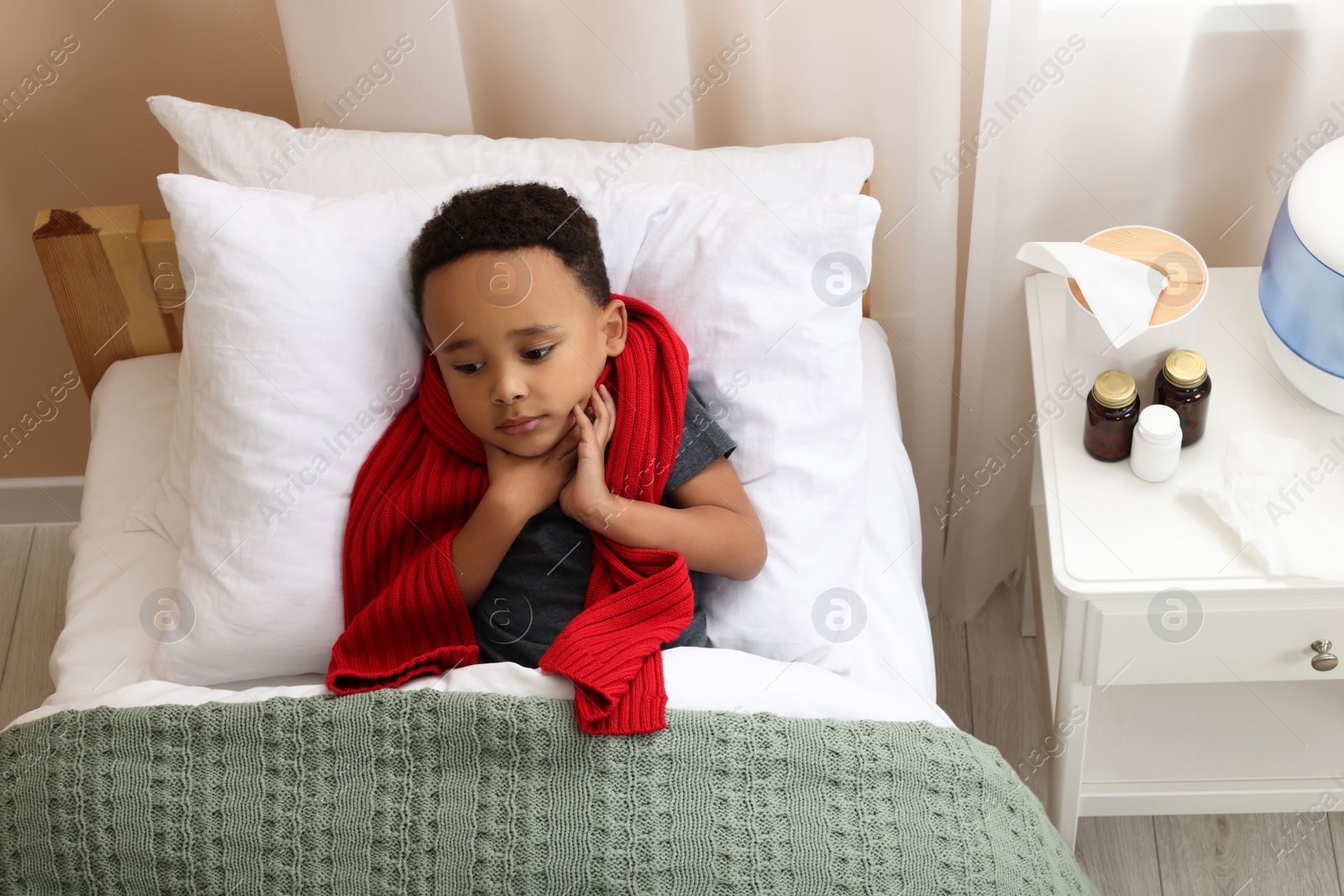 Photo of African-American boy with scarf lying in bed indoors, above view. Cold symptoms