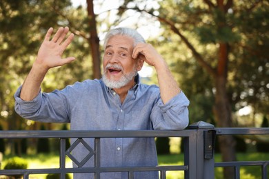 Photo of Neighbor greeting. Happy senior man waving near fence outdoors