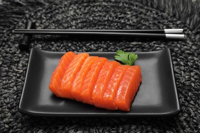 Plate of tasty salmon slices with parsley and chopsticks on black wicker mat. Delicious sashimi dish