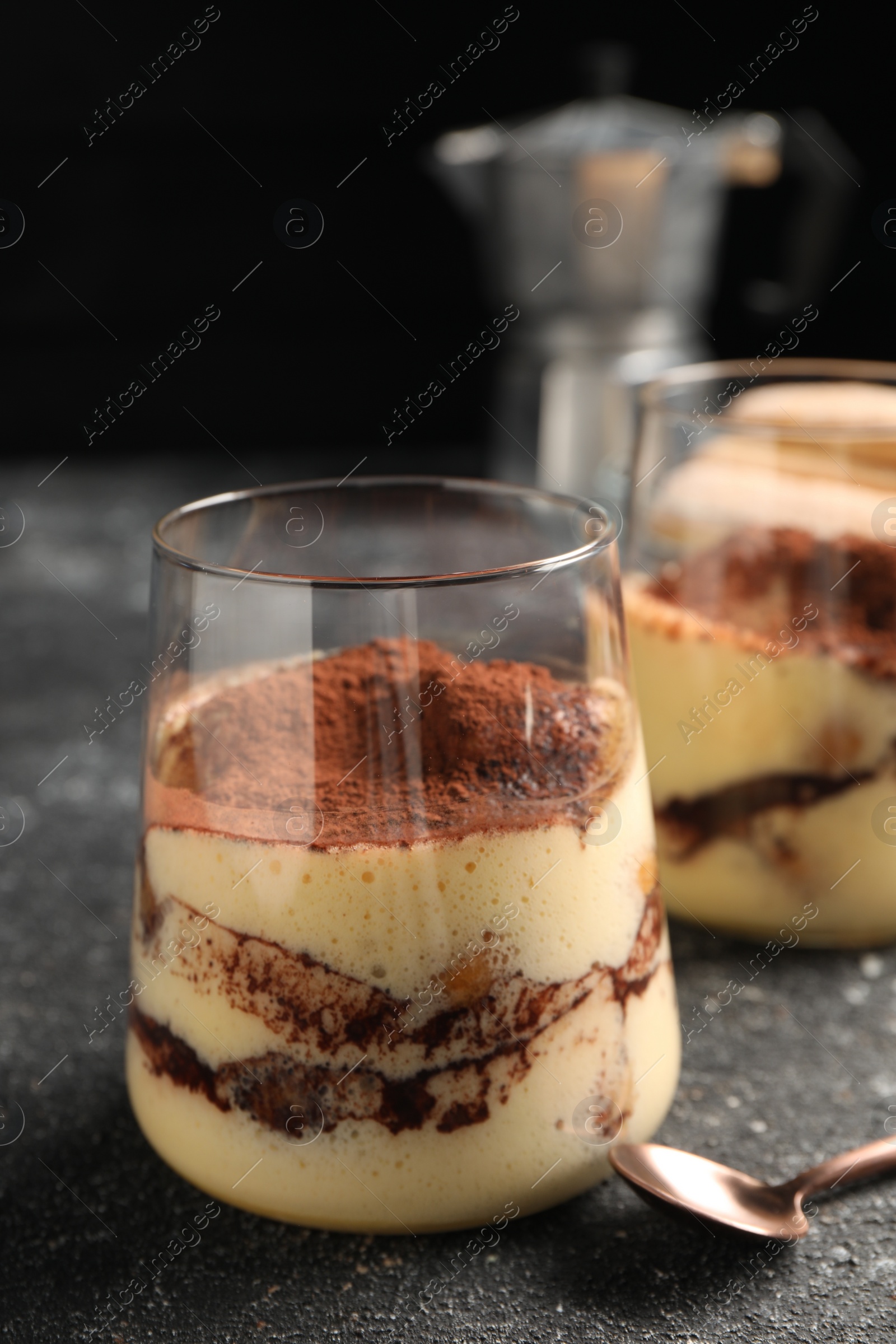 Photo of Delicious tiramisu in glasses and spoon on grey textured table, closeup