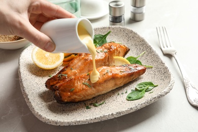 Woman pouring sauce onto tasty cooked salmon on plate, closeup