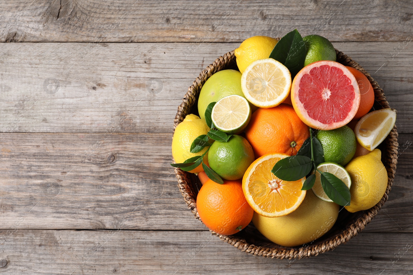 Photo of Different fresh citrus fruits and leaves in wicker basket on wooden table, top view. Space for text