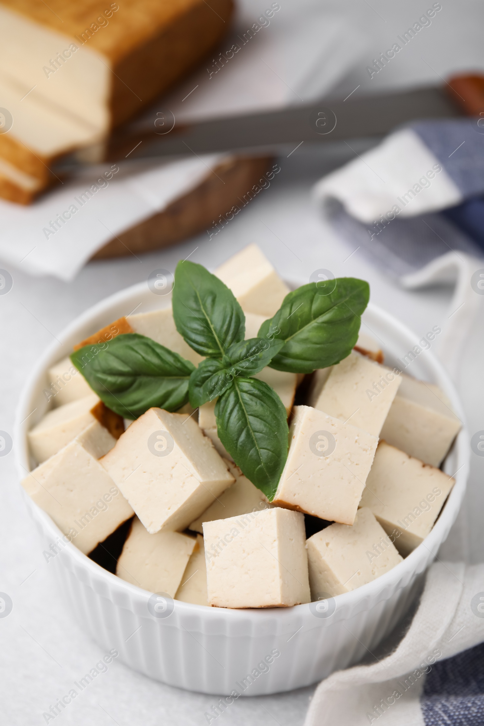 Photo of Bowl of smoked tofu cubes with basil on white table, closeup