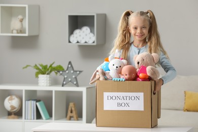 Photo of Cute little girl holding donation box with toys at home, space for text