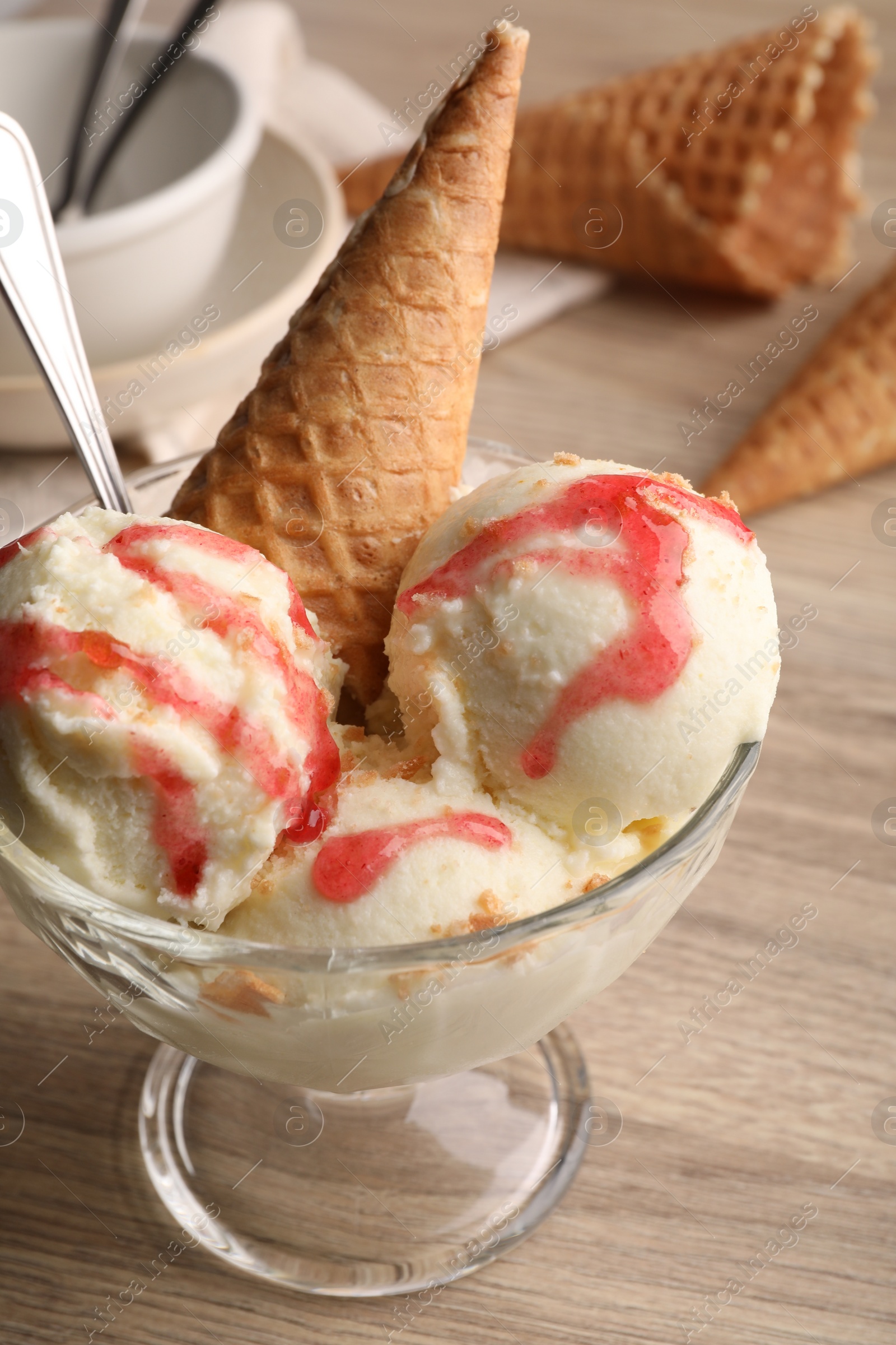 Photo of Delicious scoops of vanilla ice cream with wafer cone and strawberry topping in glass dessert bowl on wooden table, closeup