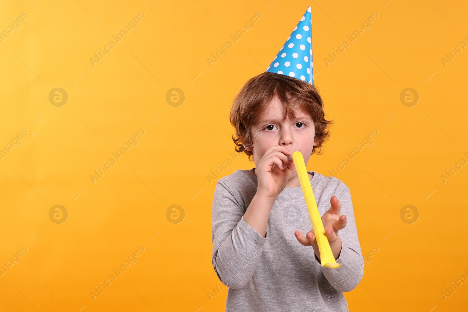 Photo of Birthday celebration. Cute little boy in party hat with blower on orange background, space for text