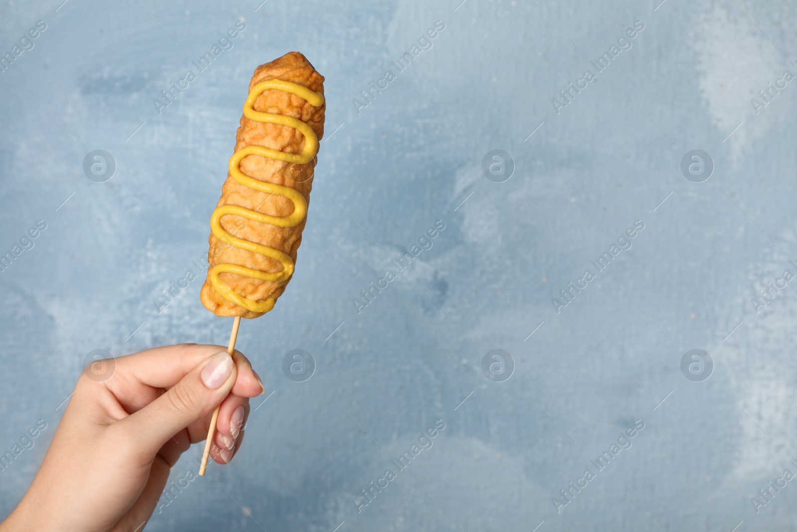 Photo of Woman holding delicious corn dog with mustard on light blue background, closeup. Space for text