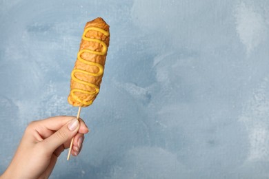 Photo of Woman holding delicious corn dog with mustard on light blue background, closeup. Space for text