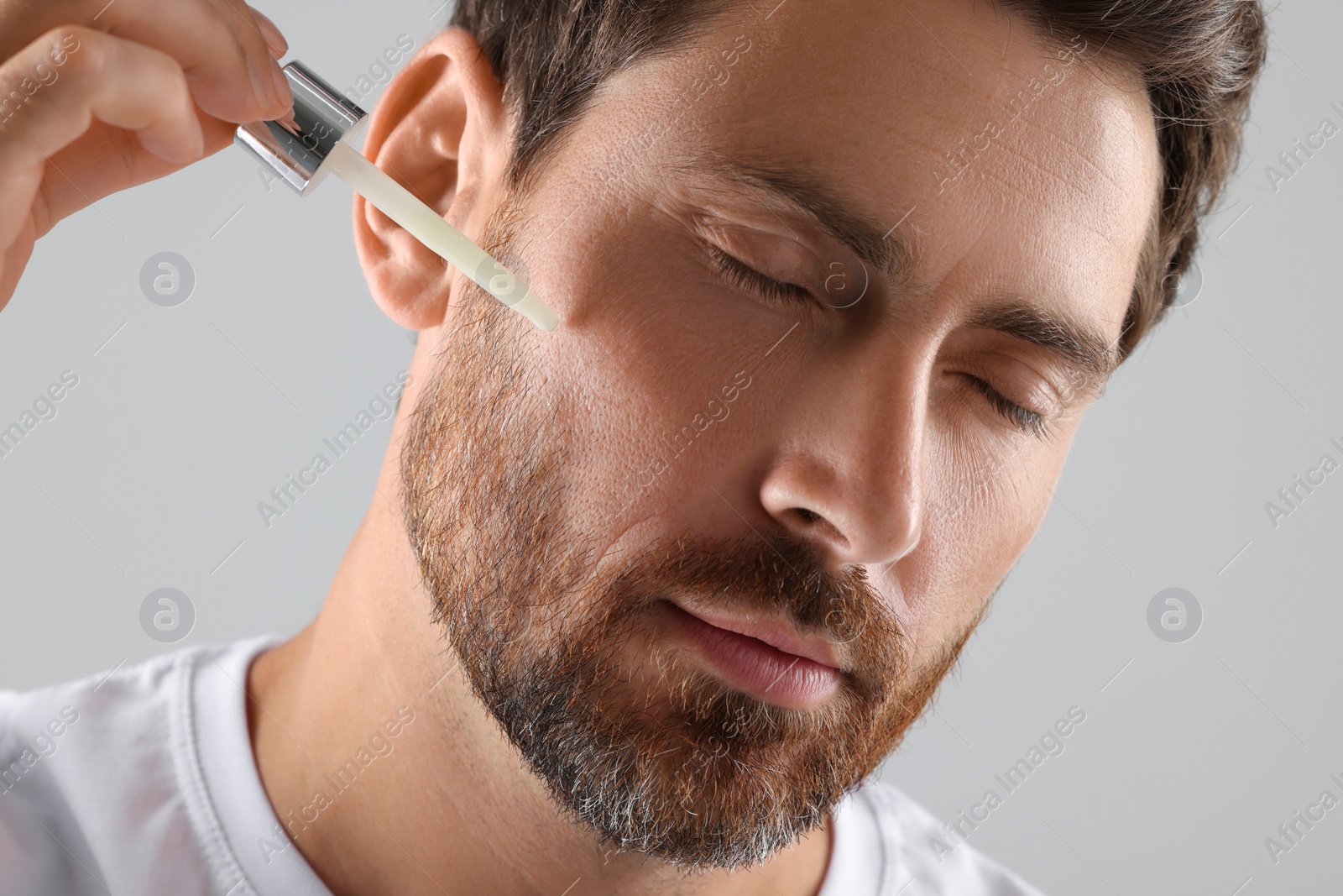 Photo of Handsome man applying cosmetic serum onto his face on light grey background, closeup