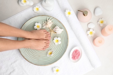 Photo of Woman putting her feet into plate with water, flowers and seashells on white towel, top view. Spa treatment