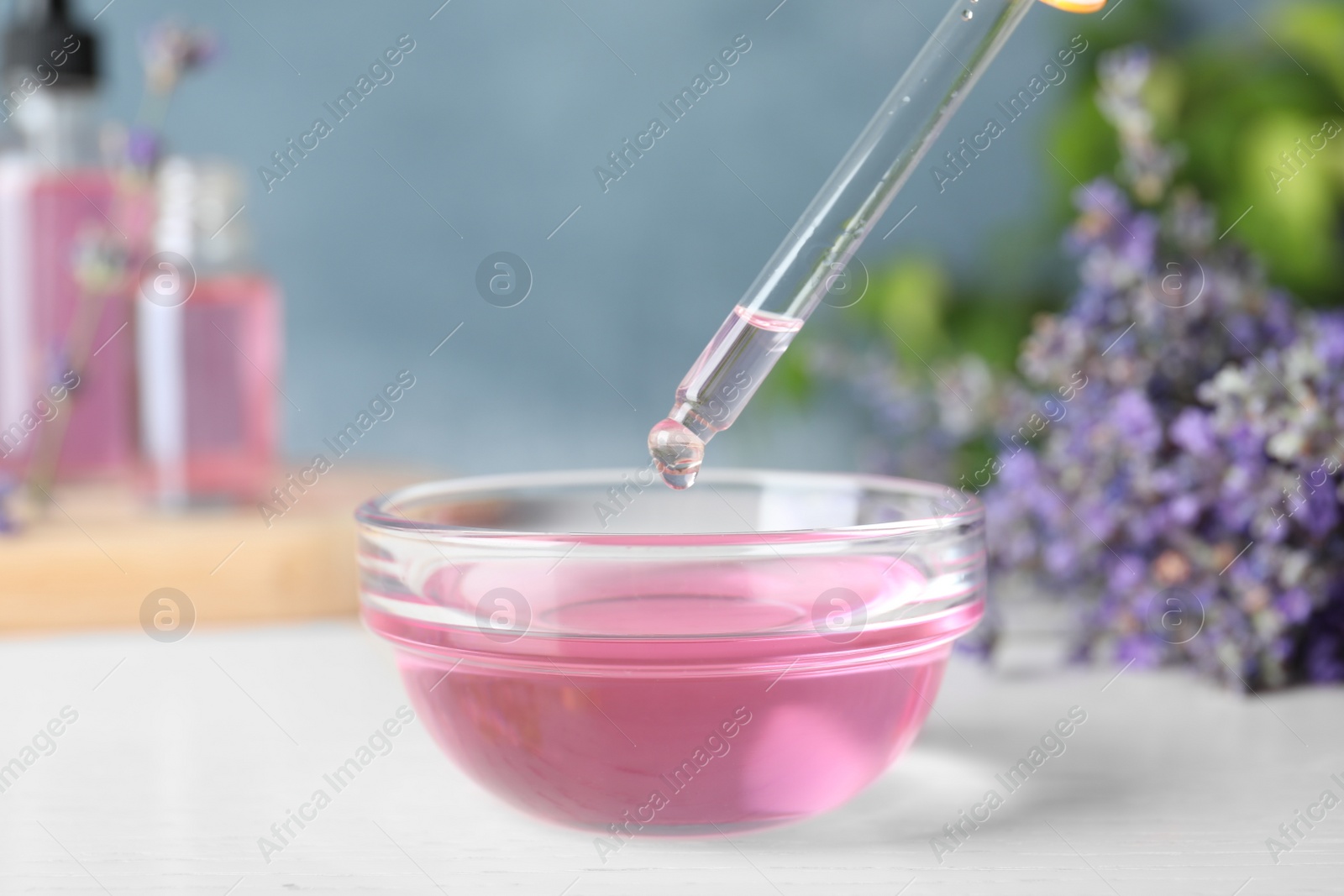 Photo of Dropper with lavender essential oil over bowl on white wooden table against blue background