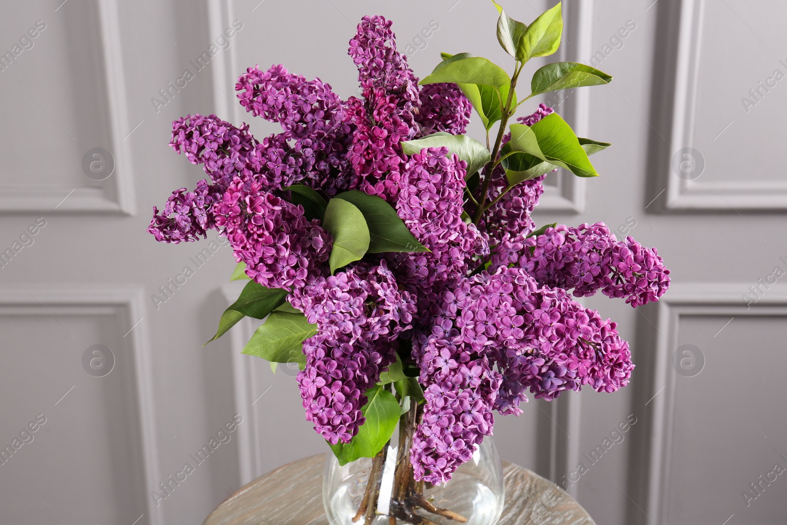 Photo of Beautiful lilac flowers in vase on wooden table near light wall