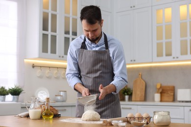 Photo of Making bread. Man sprinkling flour onto dough at wooden table in kitchen