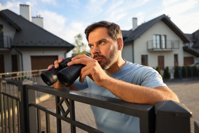 Concept of private life. Curious man with binoculars spying on neighbours over fence outdoors