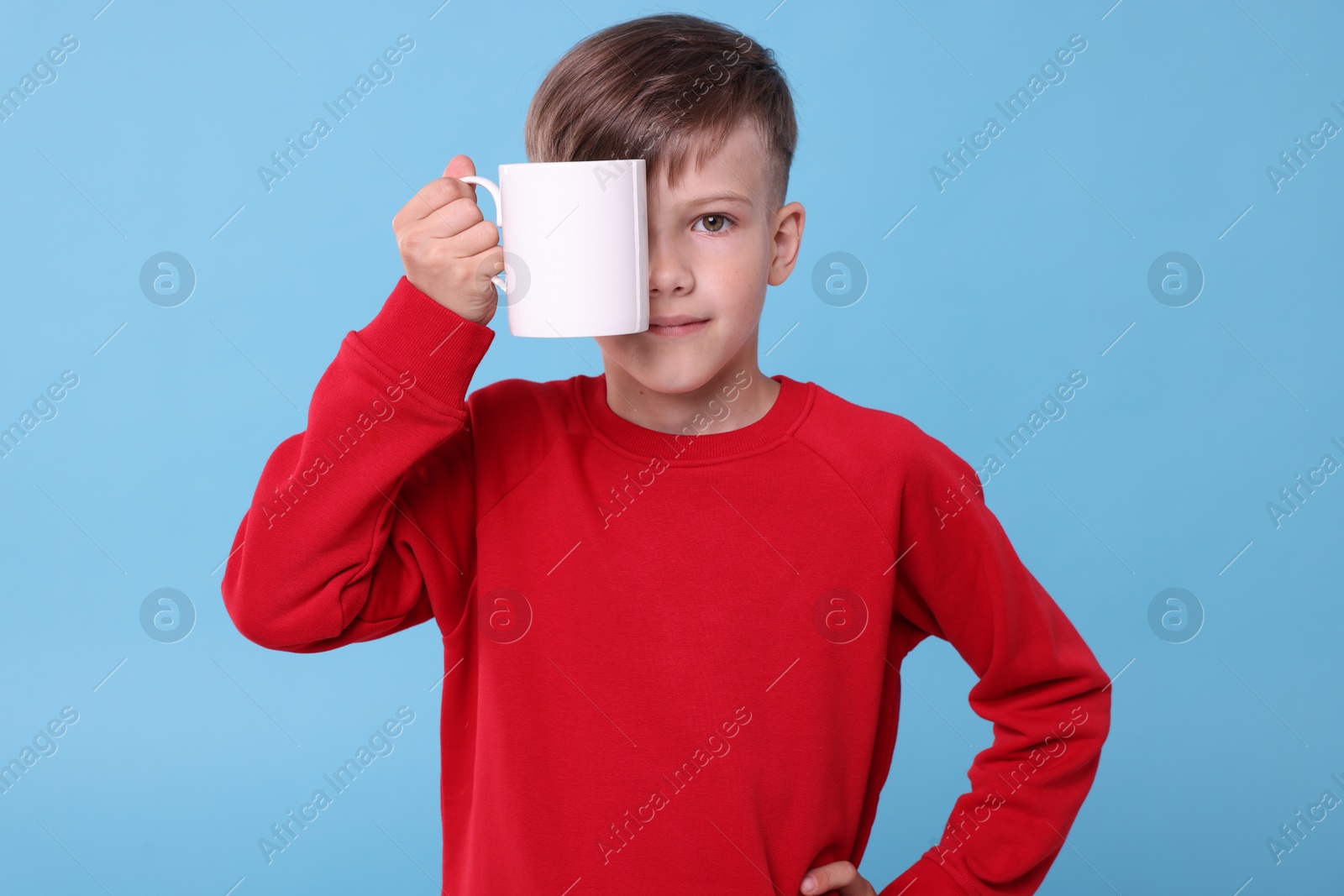 Photo of Cute boy covering eye with white ceramic mug on light blue background