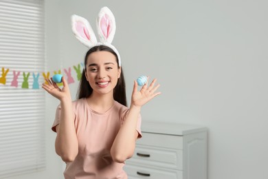 Photo of Happy woman in bunny ears headband holding painted Easter eggs indoors. Space for text