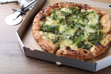 Photo of Delicious pizza with pesto, cheese and arugula in cardboard box on wooden table, closeup