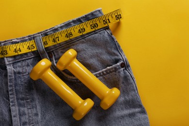 Jeans with measuring tape and dumbbells on yellow background, top view. Weight loss concept