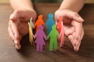 Photo of Woman protecting paper human figures on wooden table, closeup. Diversity and inclusion concept