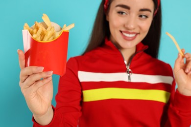 Photo of Beautiful young woman with French fries against light blue background, focus on container