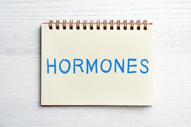 Photo of Notebook with word HORMONES on white wooden table, top view