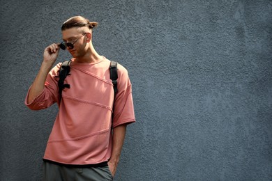 Photo of Handsome young man in stylish sunglasses and backpack near grey wall, space for text