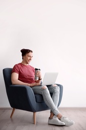 Young blogger with laptop sitting in armchair against light wall