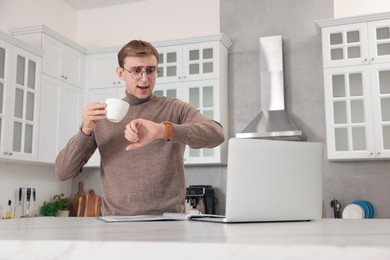 Emotional young man checking time while drinking coffee in kitchen. Being late