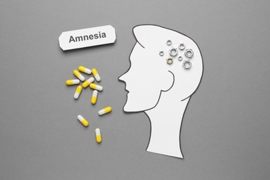 Photo of Word Amnesia, pills and human head cutout with metal nuts as brain on grey background, flat lay