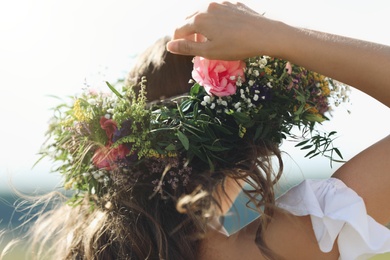 Young woman wearing wreath made of beautiful flowers outdoors on sunny day, closeup