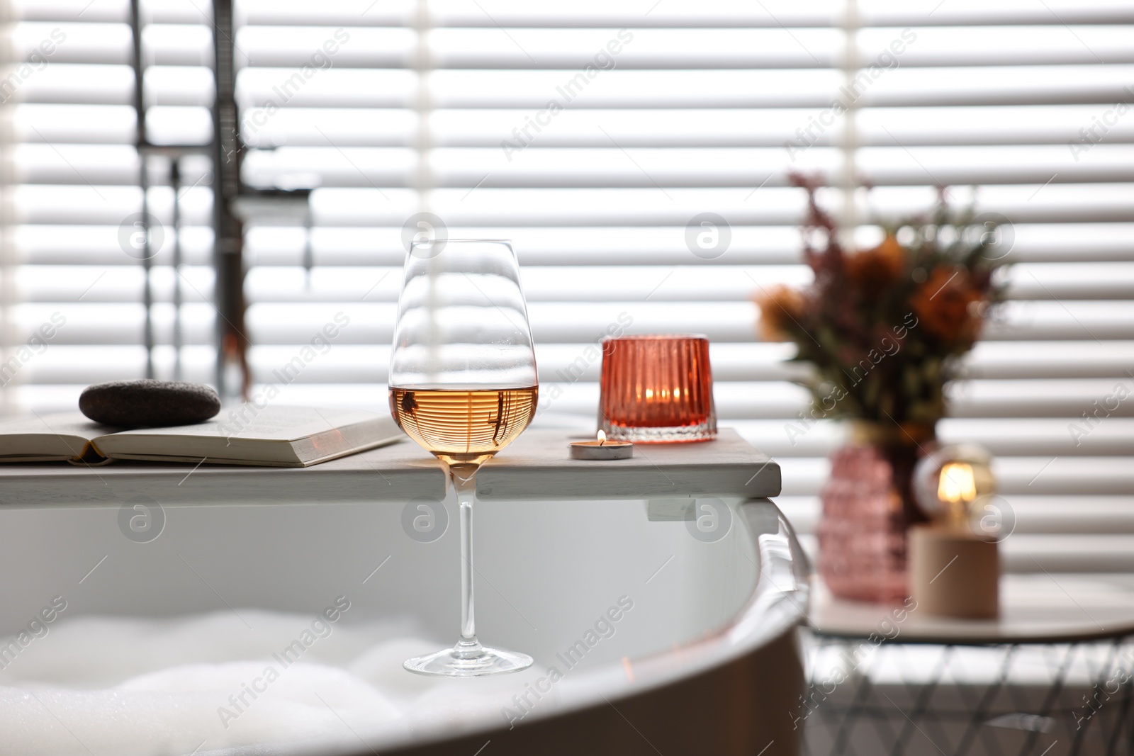 Photo of White wooden tray with glass of rose wine, book and burning candles on bathtub in bathroom