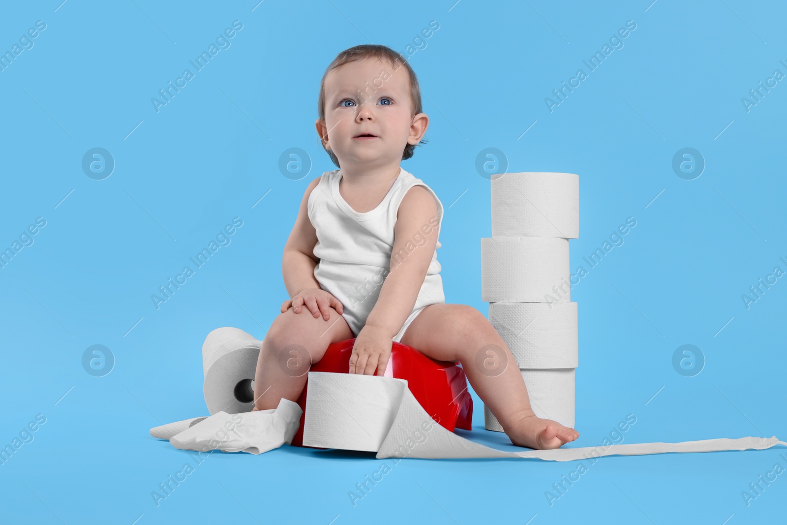Photo of Little child sitting on baby potty and stack of toilet paper rolls against light blue background