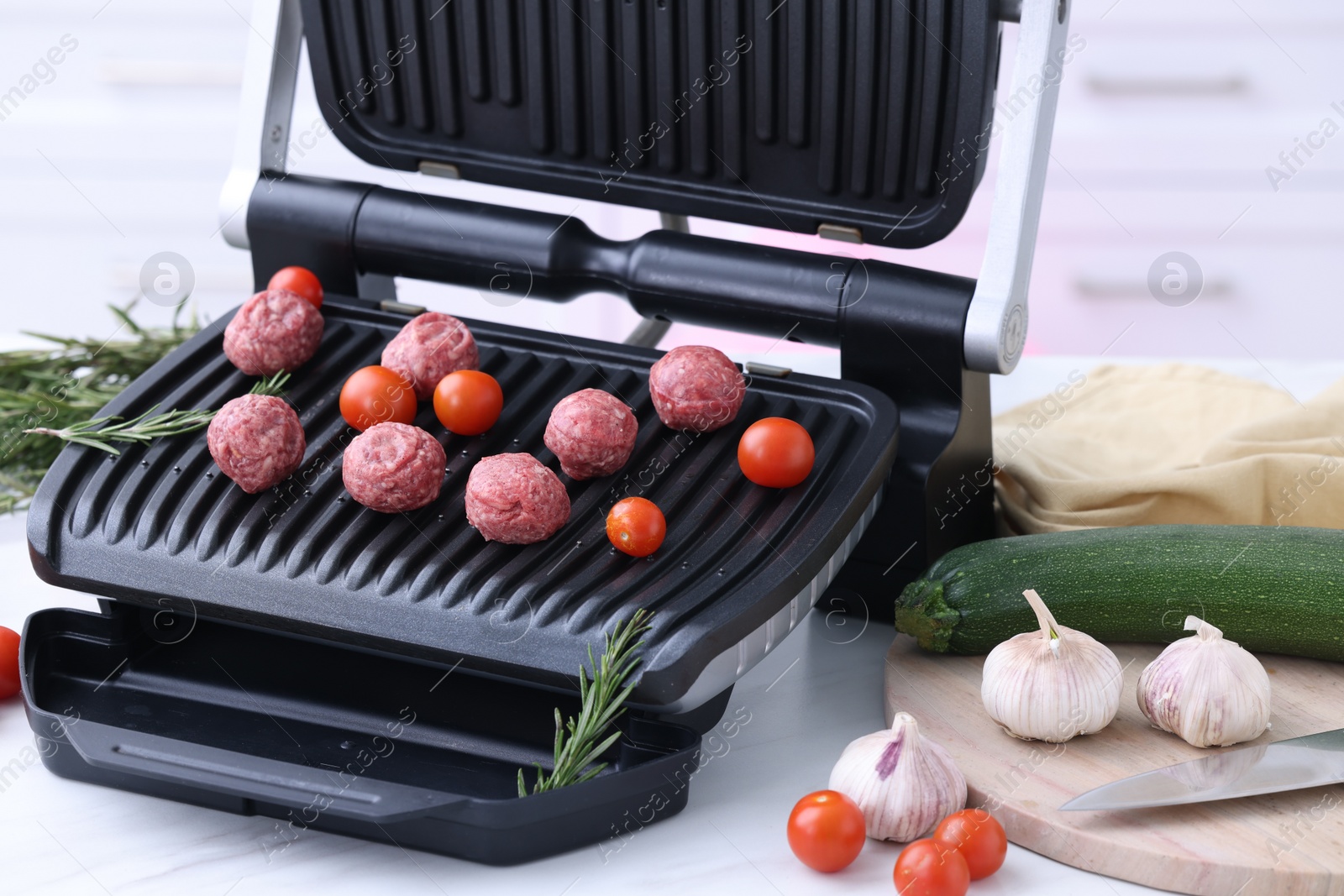 Photo of Electric grill with meatballs, tomatoes and vegetables on white table in kitchen