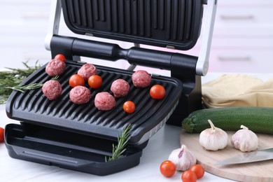 Photo of Electric grill with meatballs, tomatoes and vegetables on white table in kitchen