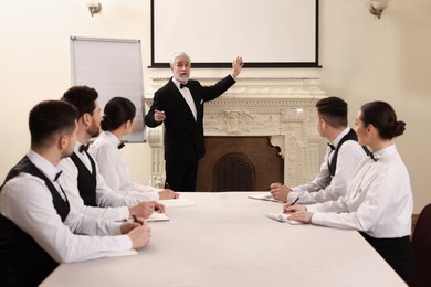 Senior man in formal suit teaching trainees indoors. Professional butler courses