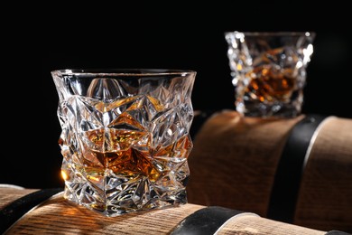 Photo of Glasses of tasty whiskey on wooden barrels against black background, closeup