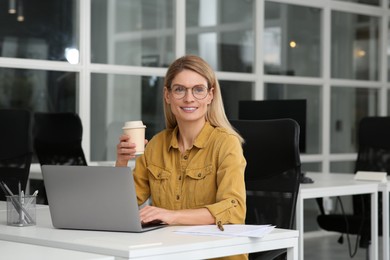 Photo of Woman with cup of coffee working on laptop at white desk in office