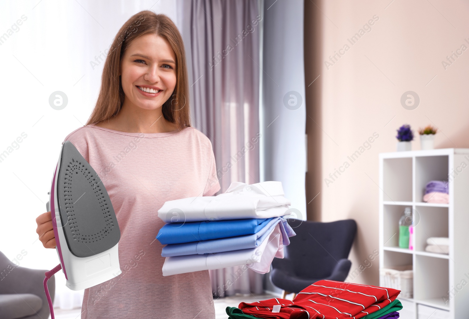 Photo of Young woman with folded clothes and iron at home. Space for text