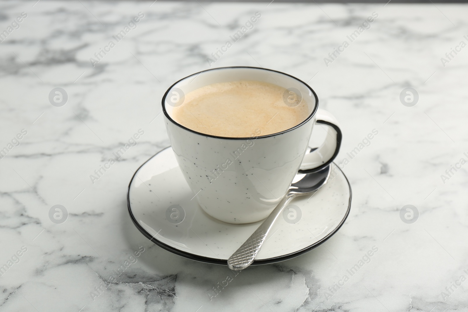 Photo of Tasty cappuccino in cup, spoon and saucer on white marble table, closeup