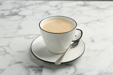 Photo of Tasty cappuccino in cup, spoon and saucer on white marble table, closeup