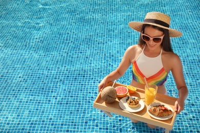 Young woman with delicious breakfast on tray in swimming pool. Space for text